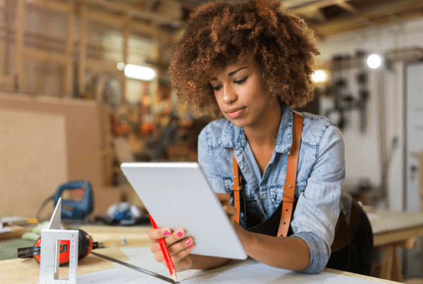 A young entrepreneur working in a workshop, reviewing plans on a tablet with a pencil in hand, surrounded by tools and materials.