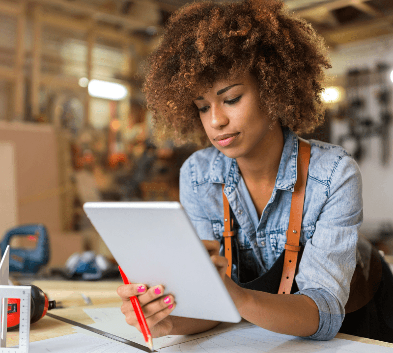 A young entrepreneur working in a workshop, reviewing plans on a tablet with a pencil in hand, surrounded by tools and materials.