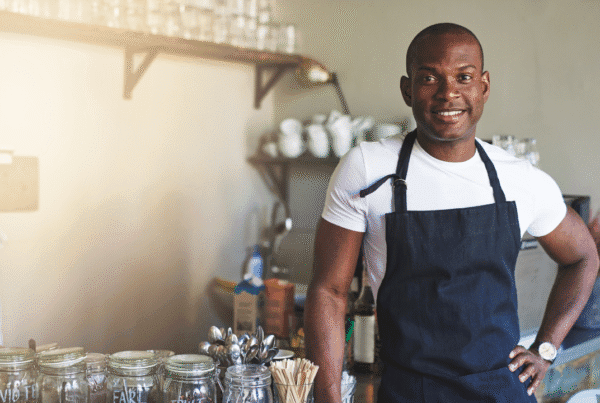 Handsome black entrepreneur stands by café counter lined with jars of tea while wearing dark colored apron.