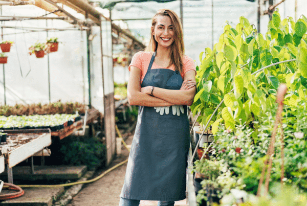 A smiling woman wearing a gardening apron stands confidently with arms crossed in a greenhouse surrounded by lush green plants and potted seedlings.