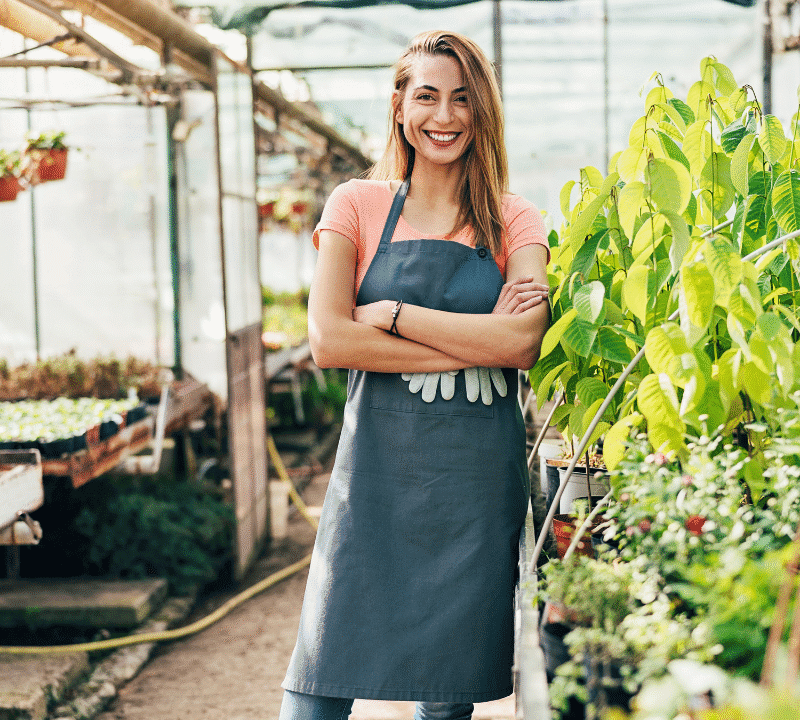 A smiling woman wearing a gardening apron stands confidently with arms crossed in a greenhouse surrounded by lush green plants and potted seedlings.