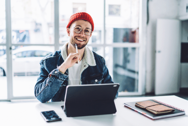 Young cheerful bearded male freelancer in glasses and warm clothes thinking happily at workplace in bright room with floor windows