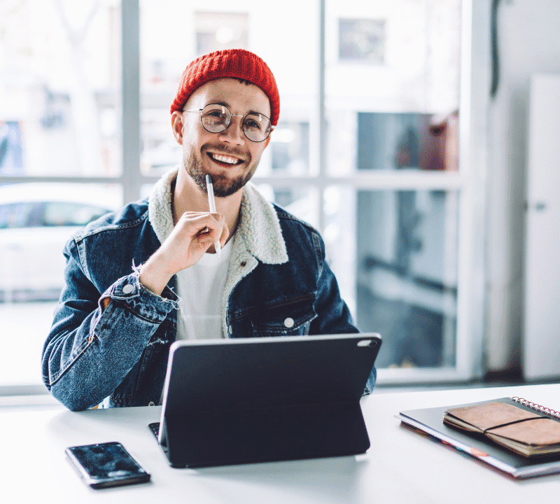 Young cheerful bearded male freelancer in glasses and warm clothes thinking happily at workplace in bright room with floor windows