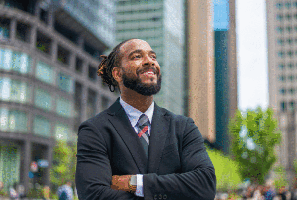 waist up portrait of a successful mid adult black entrepreneur. He is smiling and looking away with his arms crossed. He looks professional and is wearing formal business wear. Defocused city buildings in the background.