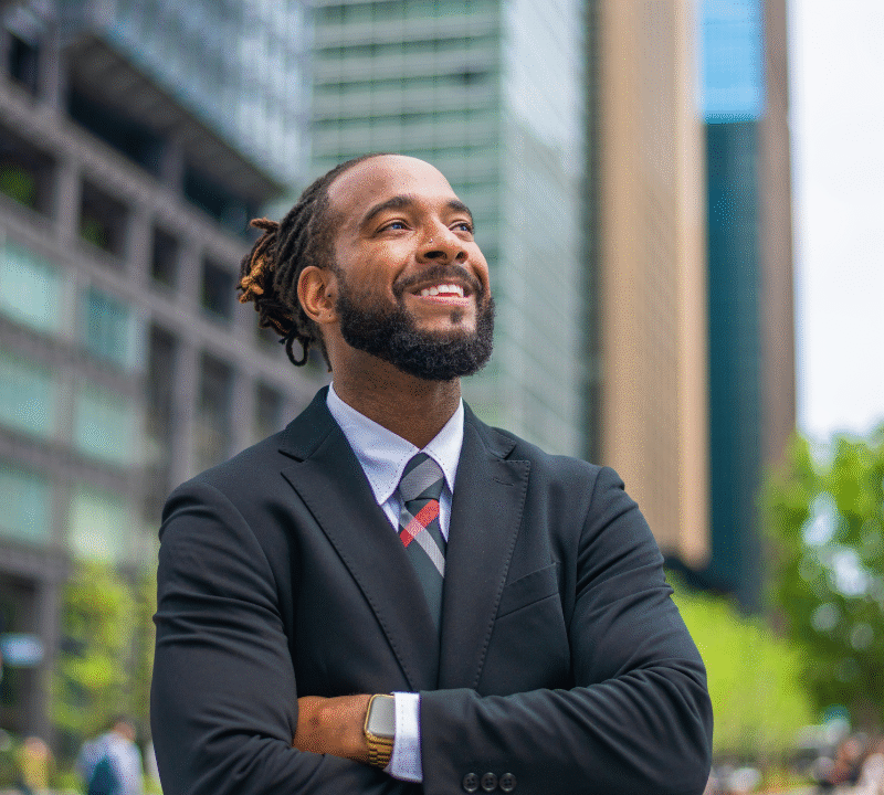 waist up portrait of a successful mid adult black entrepreneur. He is smiling and looking away with his arms crossed. He looks professional and is wearing formal business wear. Defocused city buildings in the background.