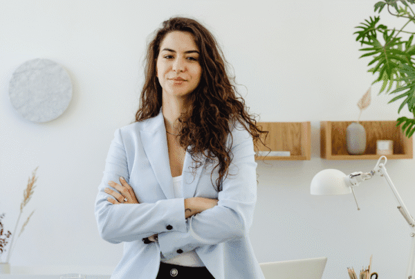 Confident businesswoman with long curly hair standing with arms crossed in a bright modern office, wearing a light blue blazer.