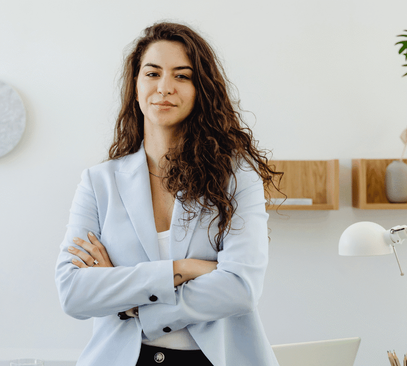 Confident businesswoman with long curly hair standing with arms crossed in a bright modern office, wearing a light blue blazer.
