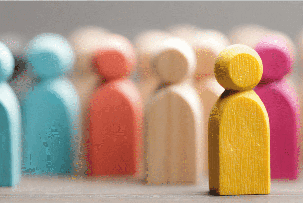 Close-up of wooden figurines painted in different colours, with a yellow figure in sharp focus standing out from a blurred background of other figures, symbolizing individuality and standing out from the crowd.