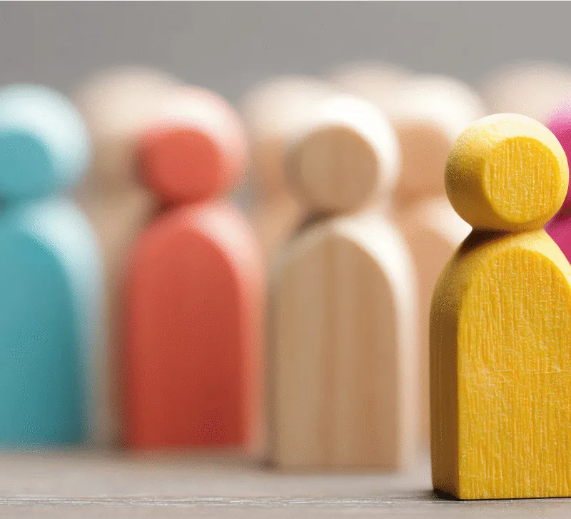 Close-up of wooden figurines painted in different colours, with a yellow figure in sharp focus standing out from a blurred background of other figures, symbolizing individuality and standing out from the crowd.