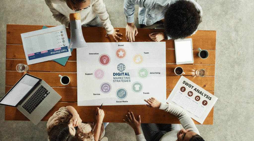 “Overhead view of a group of people gathered around a wooden table working on a digital marketing strategy. A large paper in the center displays icons and topics like brand, advertising, and social media. Laptops, coffee, and documents including a SWOT analysis are spread across the table.