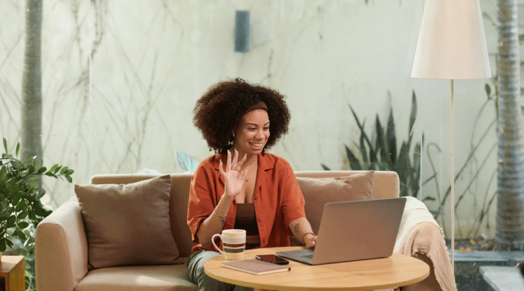 Smiling woman sitting on a couch, waving at her laptop during a video call with a coffee mug and notebook on the table