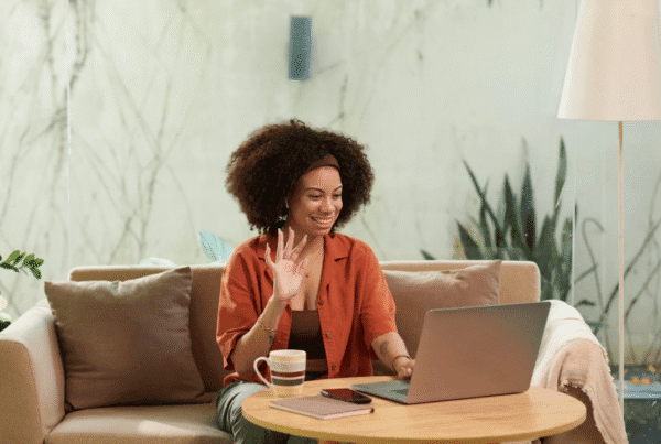 Smiling woman sitting on a couch, waving at her laptop during a video call with a coffee mug and notebook on the table