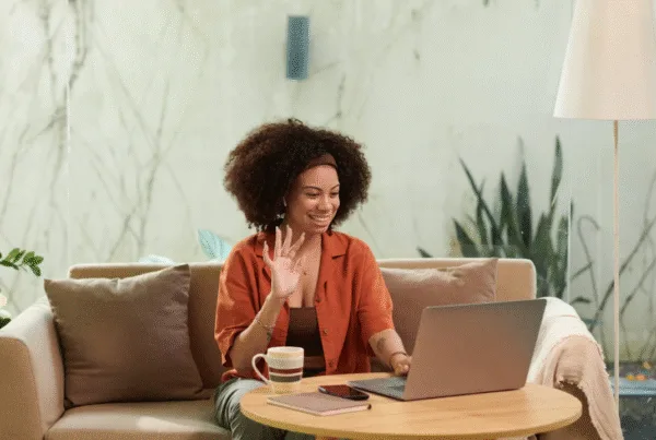 Smiling woman sitting on a couch, waving at her laptop during a video call with a coffee mug and notebook on the table