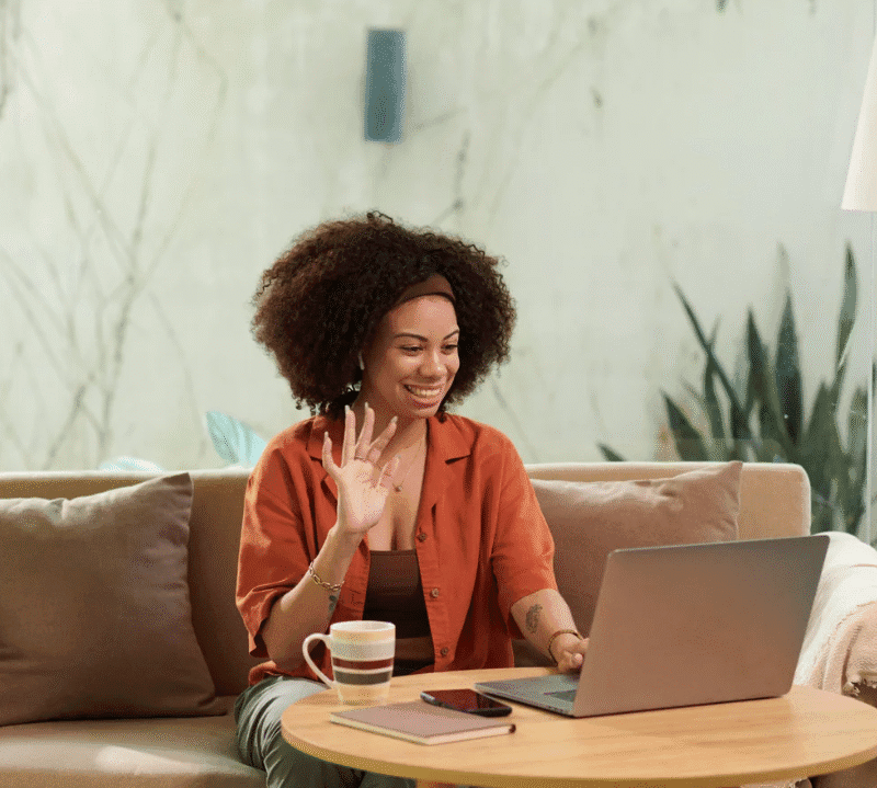 Smiling woman sitting on a couch, waving at her laptop during a video call with a coffee mug and notebook on the table