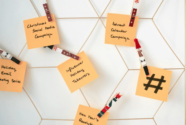 “Orange sticky notes clipped to a gold wire board with holiday-themed clothespins, displaying handwritten marketing ideas like ‘Christmas Social Media Campaign,’ ‘Holiday Email Marketing Series,’ and a hashtag symbol.”