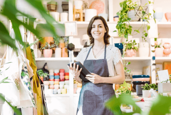Small business owner standing in a shop, holding a tablet, surrounded by products and plants.