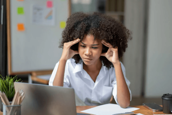 Business owner sitting at a desk looking stressed while working on a laptop, holding her temples as she reviews paperwork.