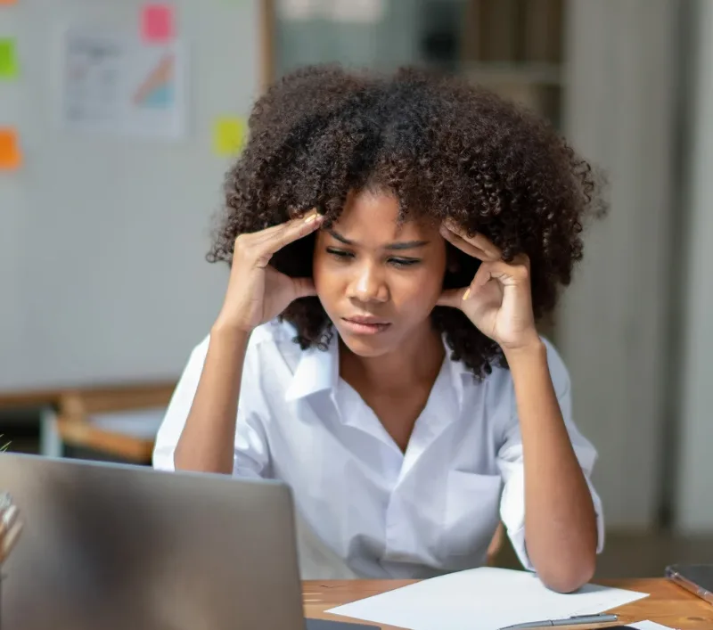 Business owner sitting at a desk looking stressed while working on a laptop, holding her temples as she reviews paperwork.