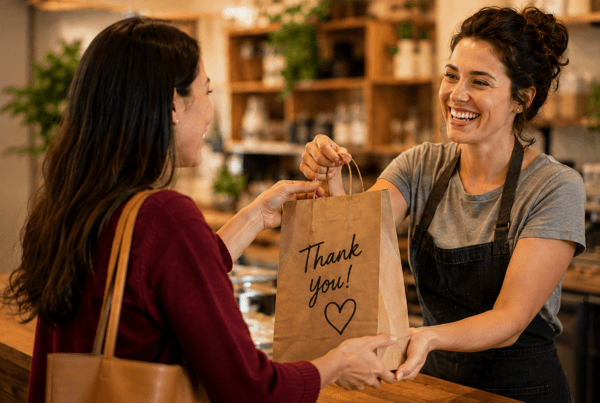 A smiling small business owner hands a customer a shopping bag across a counter, illustrating a positive customer experience, trust, and the value of building lasting customer relationships.